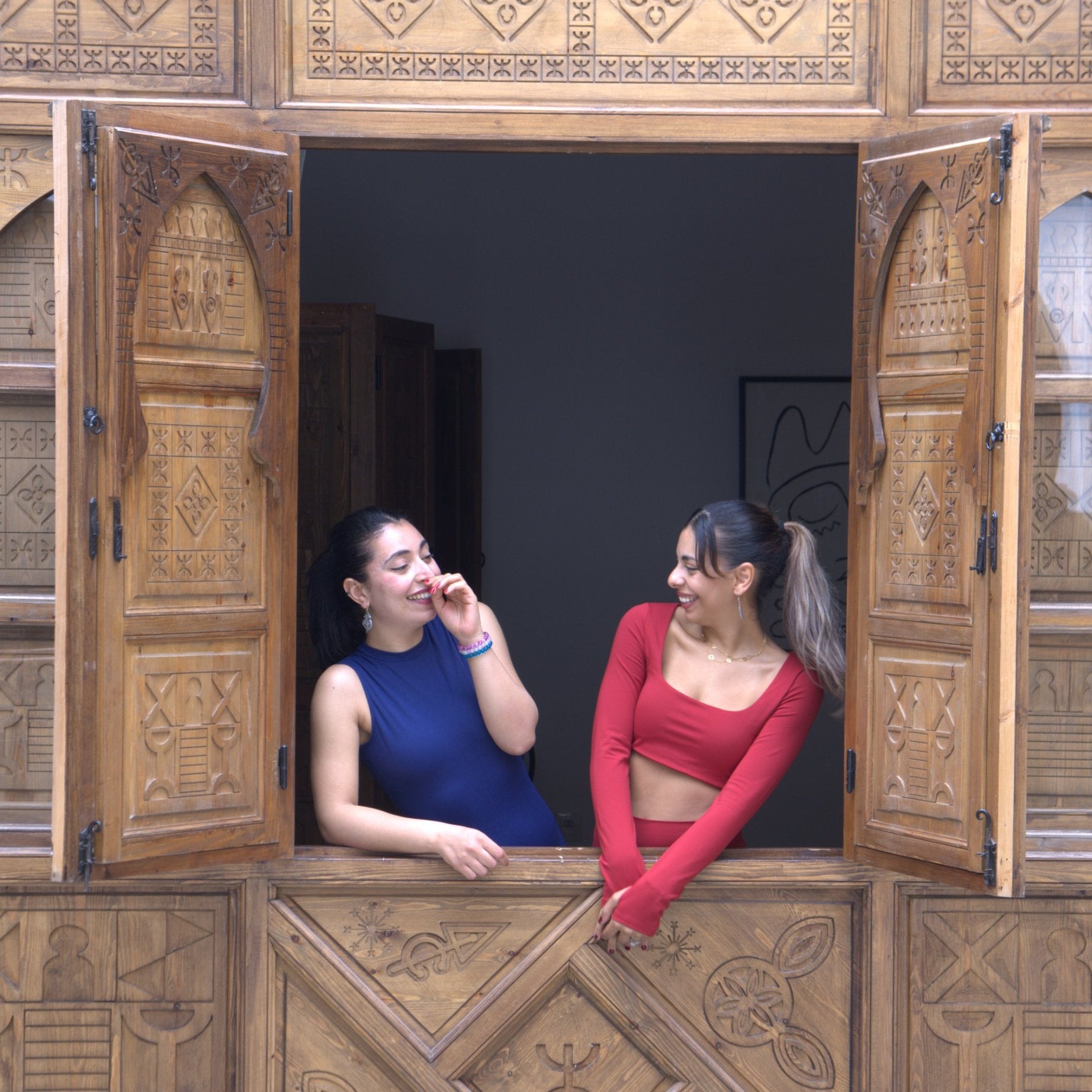 Two women leaning out of an ornate carved wooden window. One woman wears a sleeveless blue top and the other wears a red long-sleeve crop top, set against a highly detailed brown wooden screen.