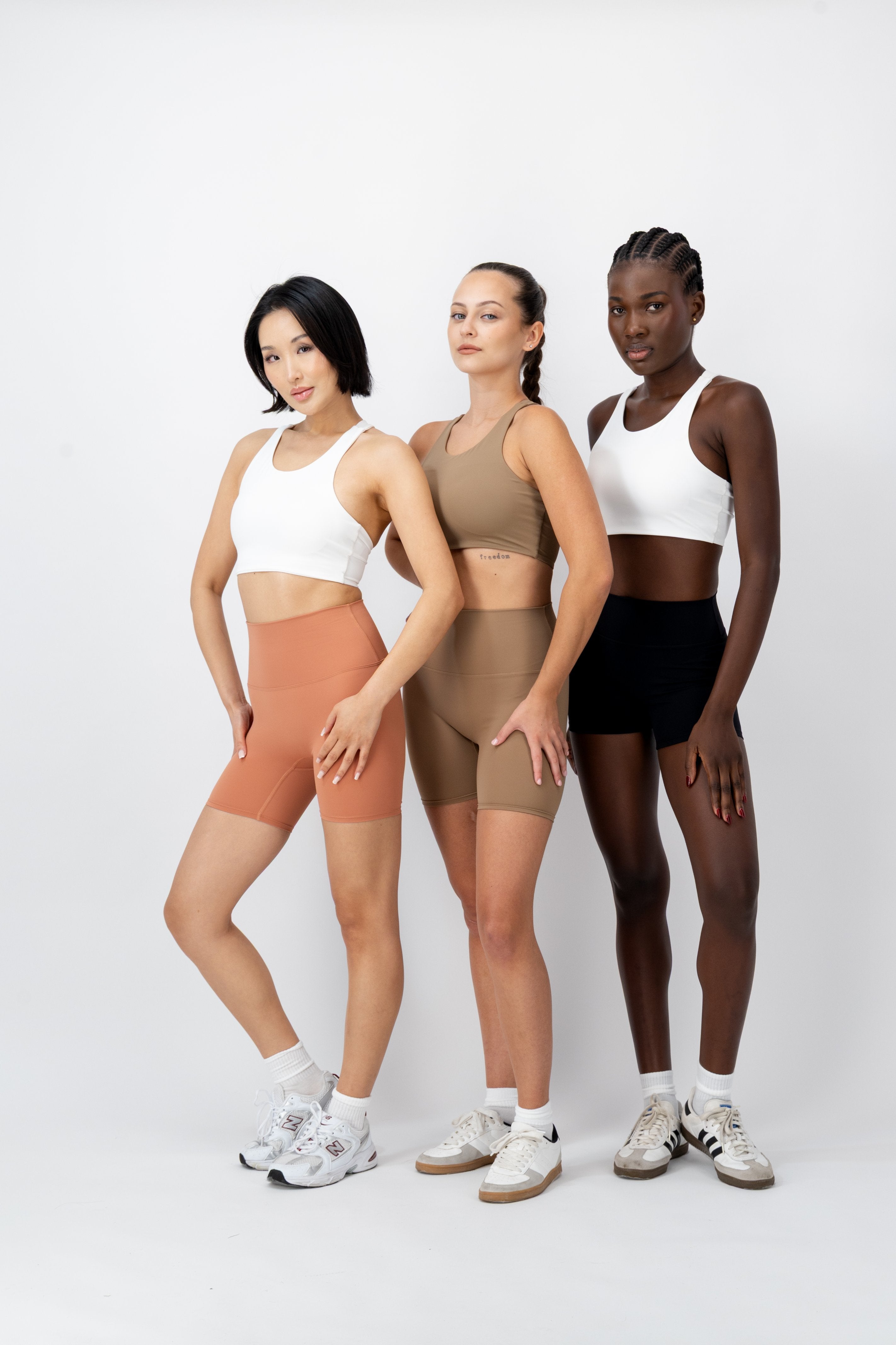 Three women modeling matching athletic wear in different colors: white sports bra and peach shorts, beige sports bra and taupe shorts, and white sports bra and black shorts, standing against a light gray studio background.