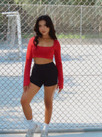 Woman in red long-sleeve top and black shorts standing on a tennis court.