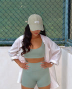 Woman wearing a light green sports bra and shorts with a white jacket and cap, standing against a chain-link fence.