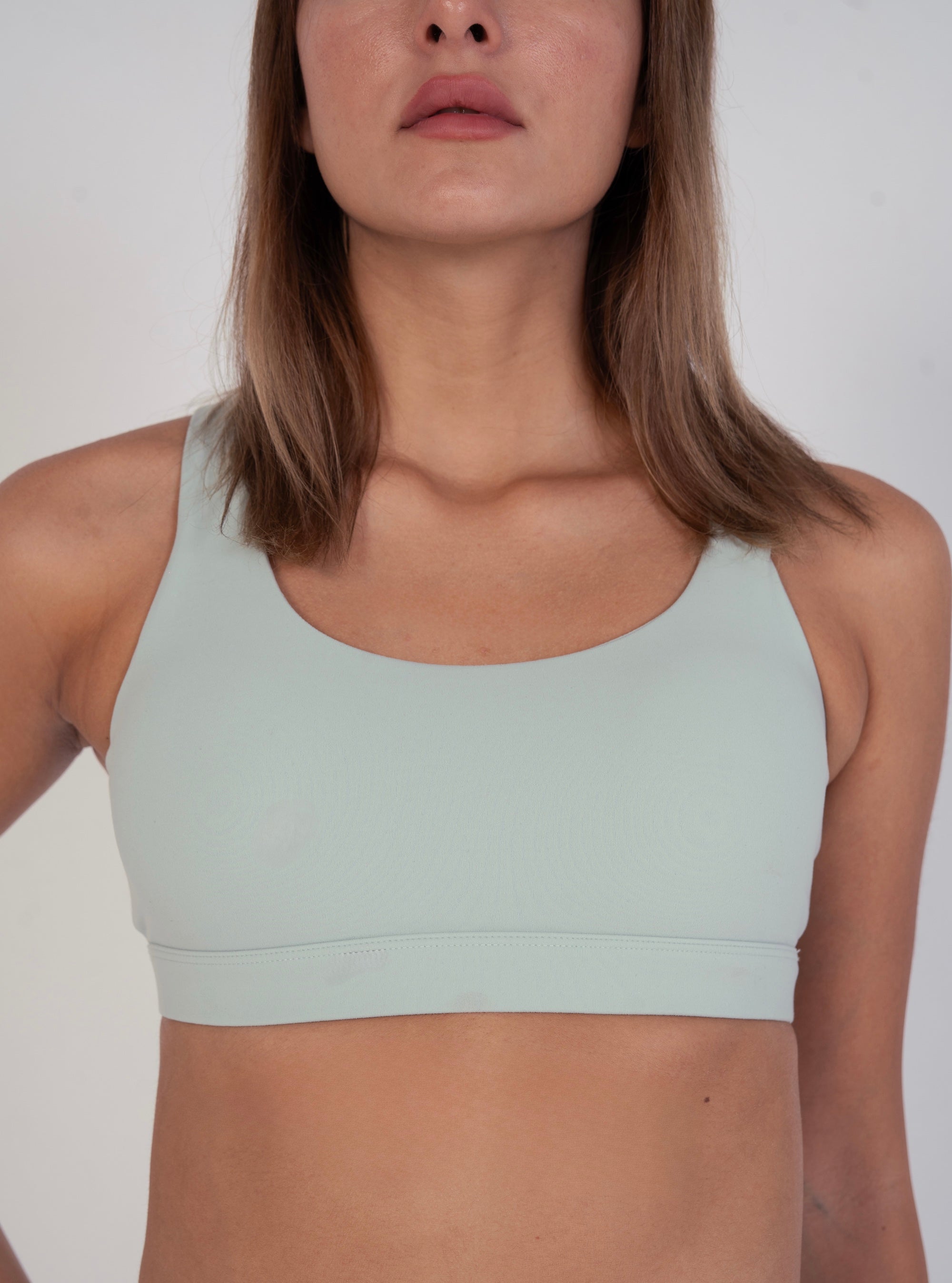 Front view of NAVA Light Sage Green sports bra, featuring a clean scoop neckline and supportive fit on model against a neutral background.

