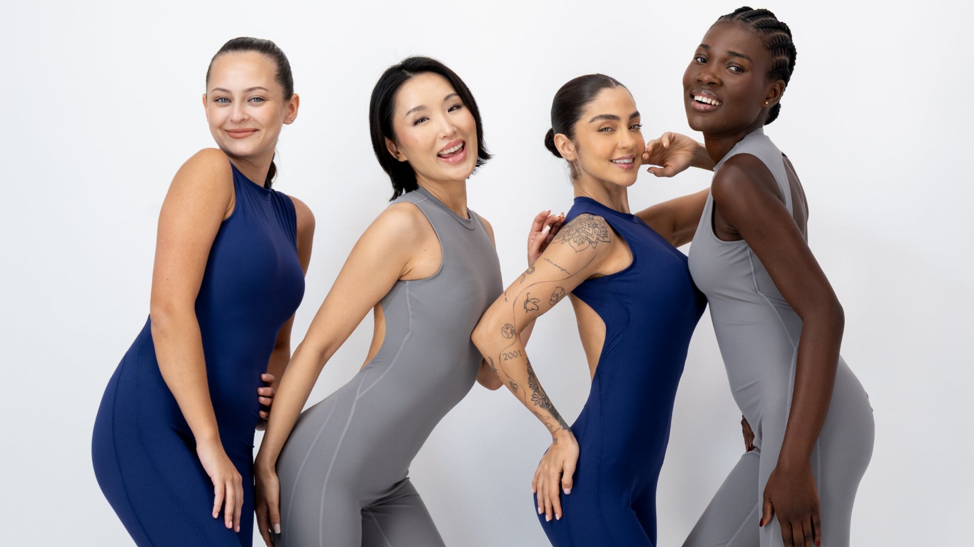 Four women posing together in matching sleeveless athletic jumpsuits in navy blue and gray against a clean white studio background.