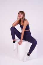 Woman wearing NAVA Sodalite Blue bra top and leggings set, sitting in a relaxed pose on a white cube in studio.